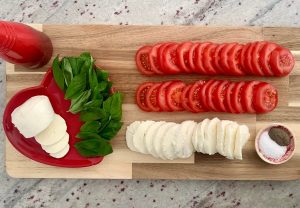 Ingredients for a Caprese Candy Cane laid out on a wooden board with two lines of sliced tomatoes, a line of sliced mozzarella, a small dish of sea salt and cracked black pepper, a bottle of balsamic vinegar and a heart-shaped red dish with basil leaves and more mozzarella.
