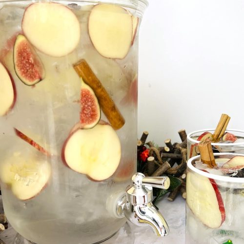 Water dispenser filled with apple slices, fig pieces and cinnamon sticks, ice and water to create infused Christmas water, with apple pieces in foreground and two garnished glasses containing infused Christmas water to the side.