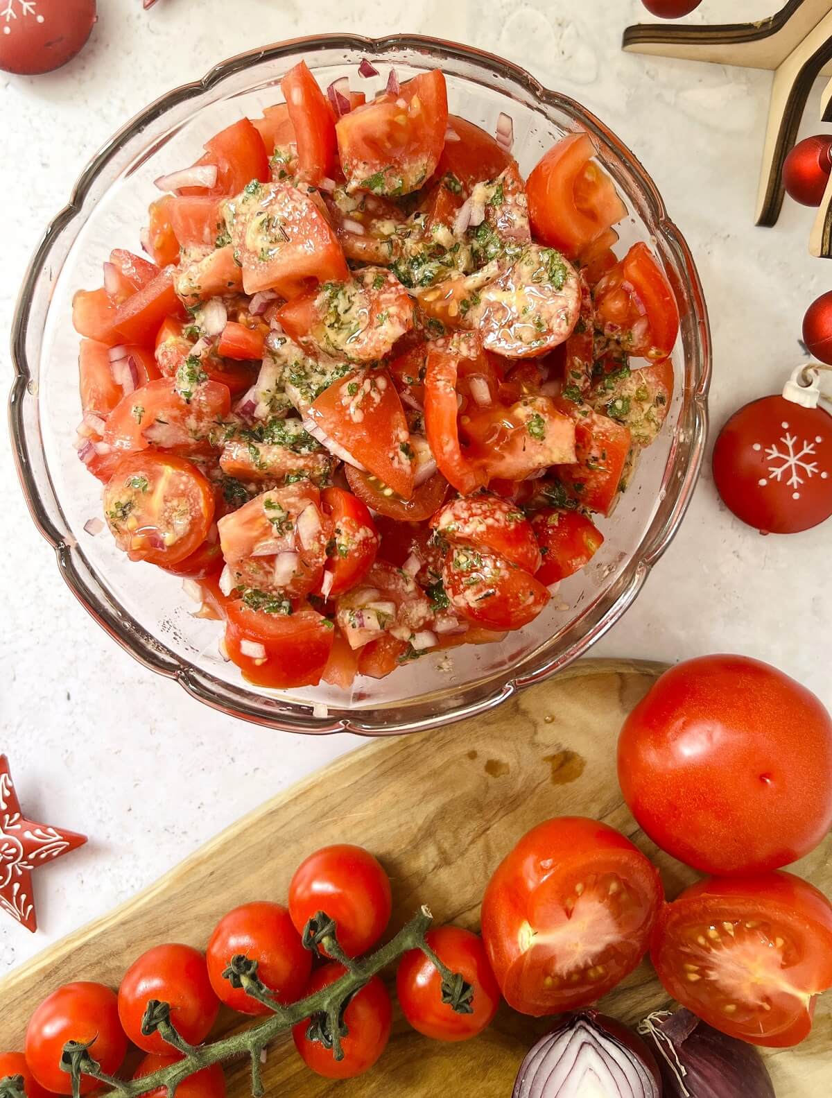 Overhead view of glass bowl containing diced red tomatoes and red onion with dressing to make a Christmas red tomato salad, with wooden board with cherry tomatoes, halved large tomato and red onion below and red metal Christmas ornaments half in shot around edge of photograph.