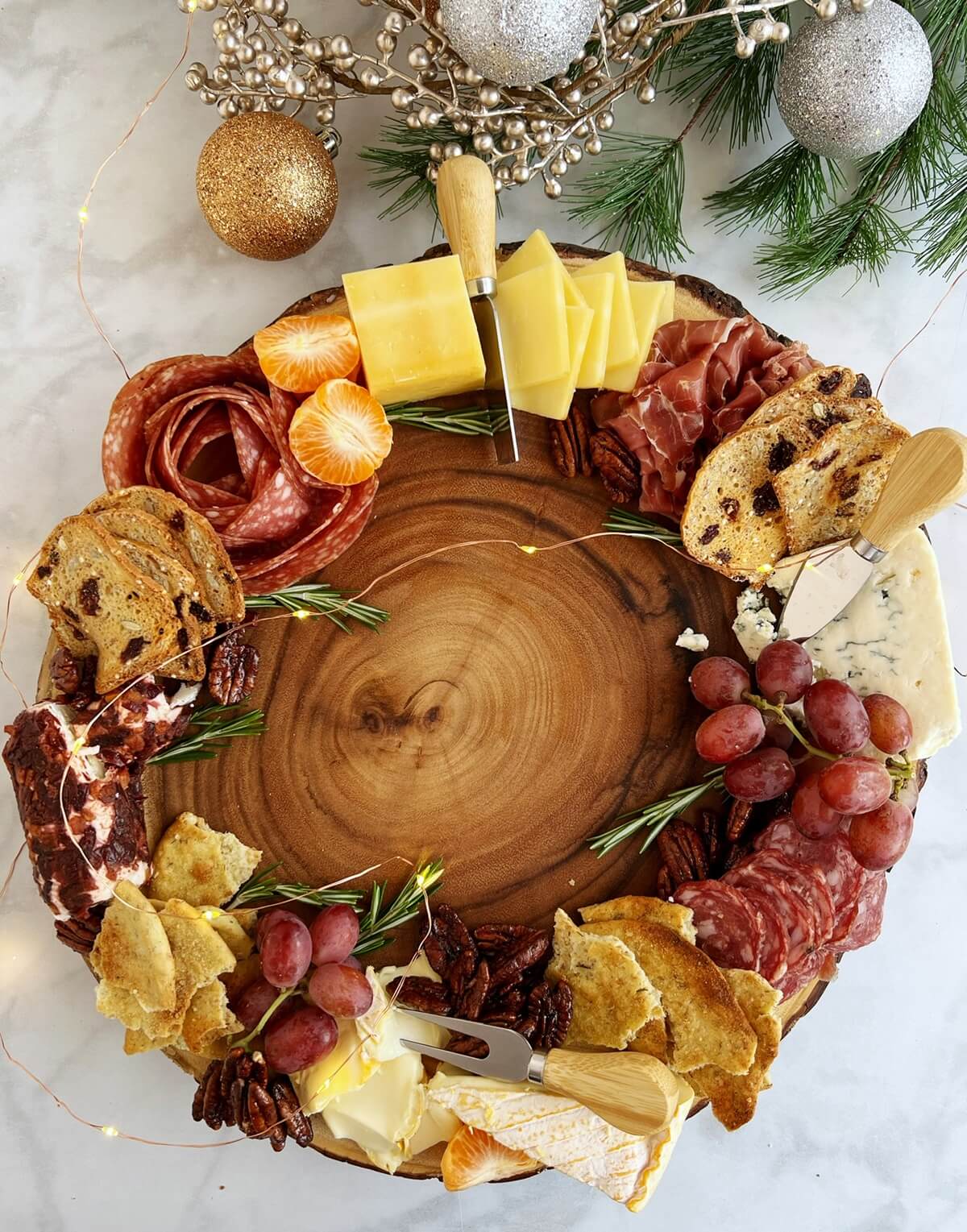 Christmas Wreath Charcuterie Board Round wooden board with charcuterie treats laid out as a wreath on top, including cheeses, salami roses, grapes, crackers, decorative rosemary and clementines with some silver and gold Christmas ornaments framing the shot at the top.