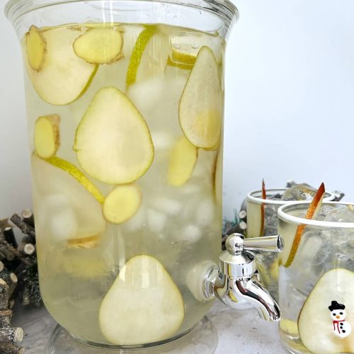 Water dispenser filled with pear and ginger slices, ice and water to create infused Christmas water, with ginger and pear slices in foreground and two glasses containing pear and ginger water to the side.