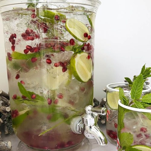 Water dispenser filled with lime slices, mint sprigs and pomegranate arils, ice and water to create infused Christmas water, with lime slices and pomegranate seeds in foreground and two glasses containing infused Christmas water with garnish to the side.