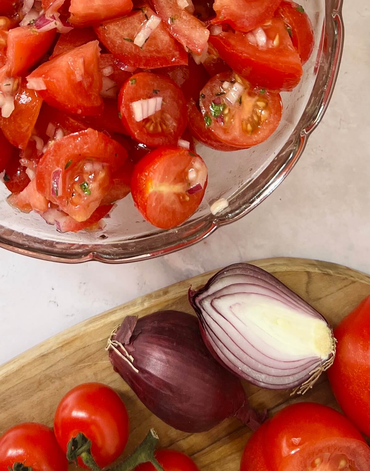 Overhead close up view of part of glass bowl containing diced red tomatoes and red onion with dressing to make a Christmas red tomato salad, with wooden board with cherry tomatoes, halved large tomato and red onion below.