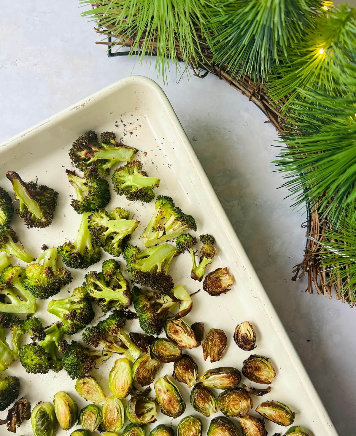Baking sheet with roasted broccoli and Brussel sprouts and fairy-lit pine in the top corner of the image.