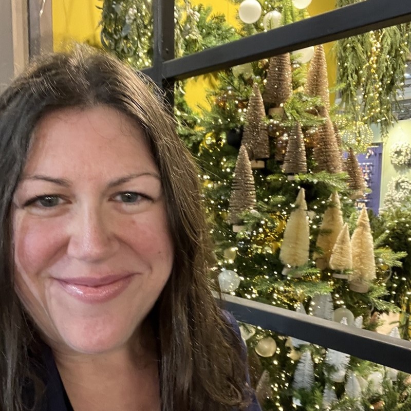 Hannah Bartlett Jolly Festive Square - Low Res Hannah Bartlett from Jolly Festive at a Christmas trade show with a display of bottlebrush Christmas trees in the background.