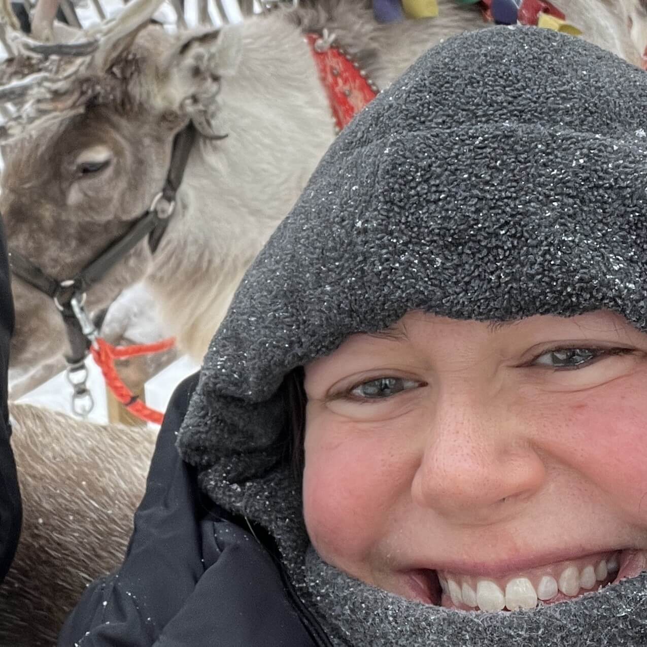 Hannah in Santa Claus Village Lapland Feb 2024 Close up of a woman (Hannah from Jolly Festive) wrapped up in a fleece balaclava in the snow with a reindeer behind her.