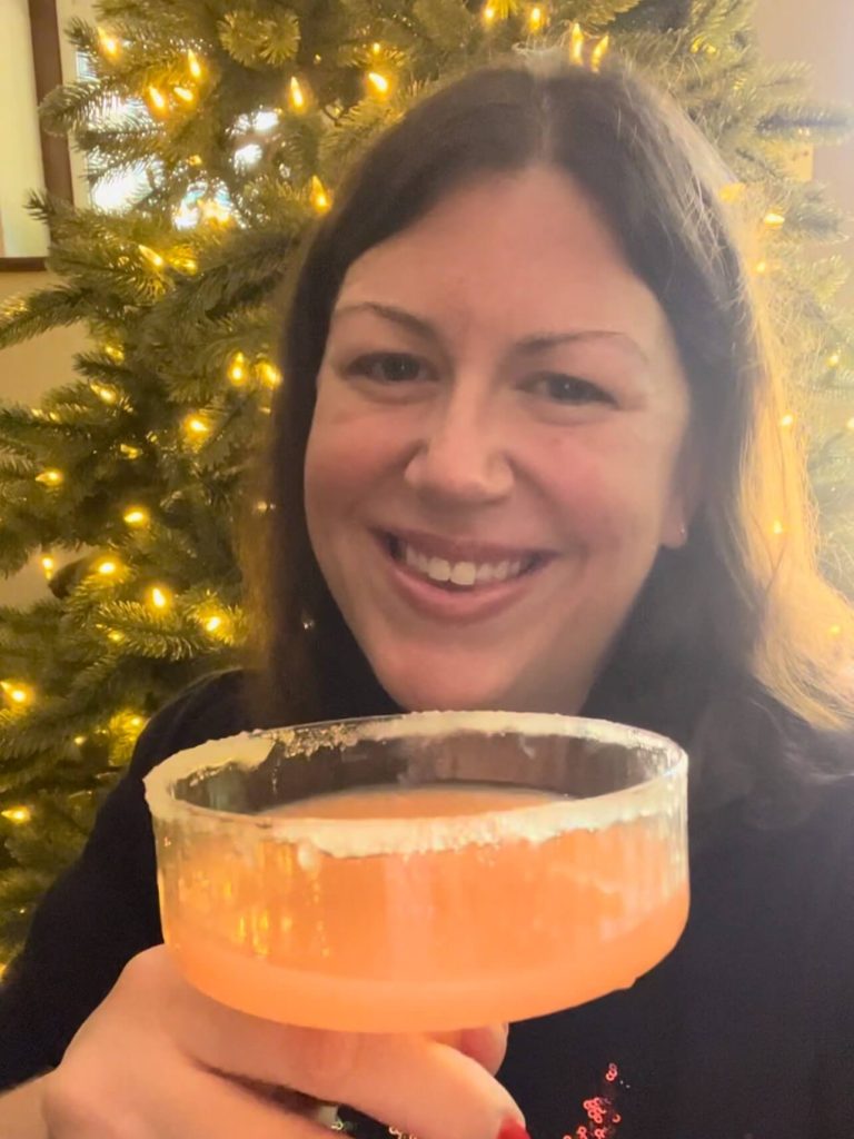 Hannah sipping homemade berry & candy cane gin Woman (Hannah from Jolly Festive) standing in front of a fairy-lit Christmas tree with a coupe of light blush coloured cocktail in her hand.