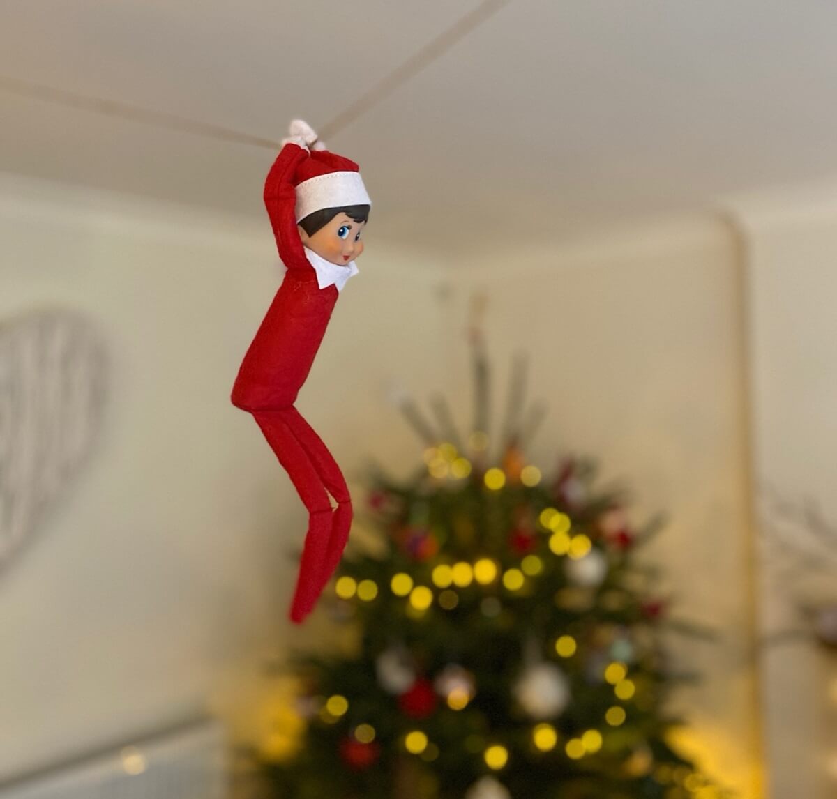 Elf Zipwires Into Room In Front Of Christmas Tree Elf on the Shelf suspended from string as if on a zipwire in front of the Christmas tree.