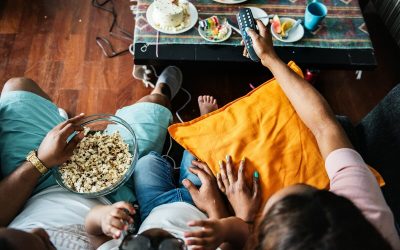 Family sitting on a couch with a bowl of popcorn, remote, cake, and fruit on the table nearby watching Christmas adverts on the television.