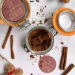 Close-up of a jar of homemade gingerbread sugar scrub with a small wooden scoop and festive cinnamon stick decorations, ideal for DIY Christmas gifts.