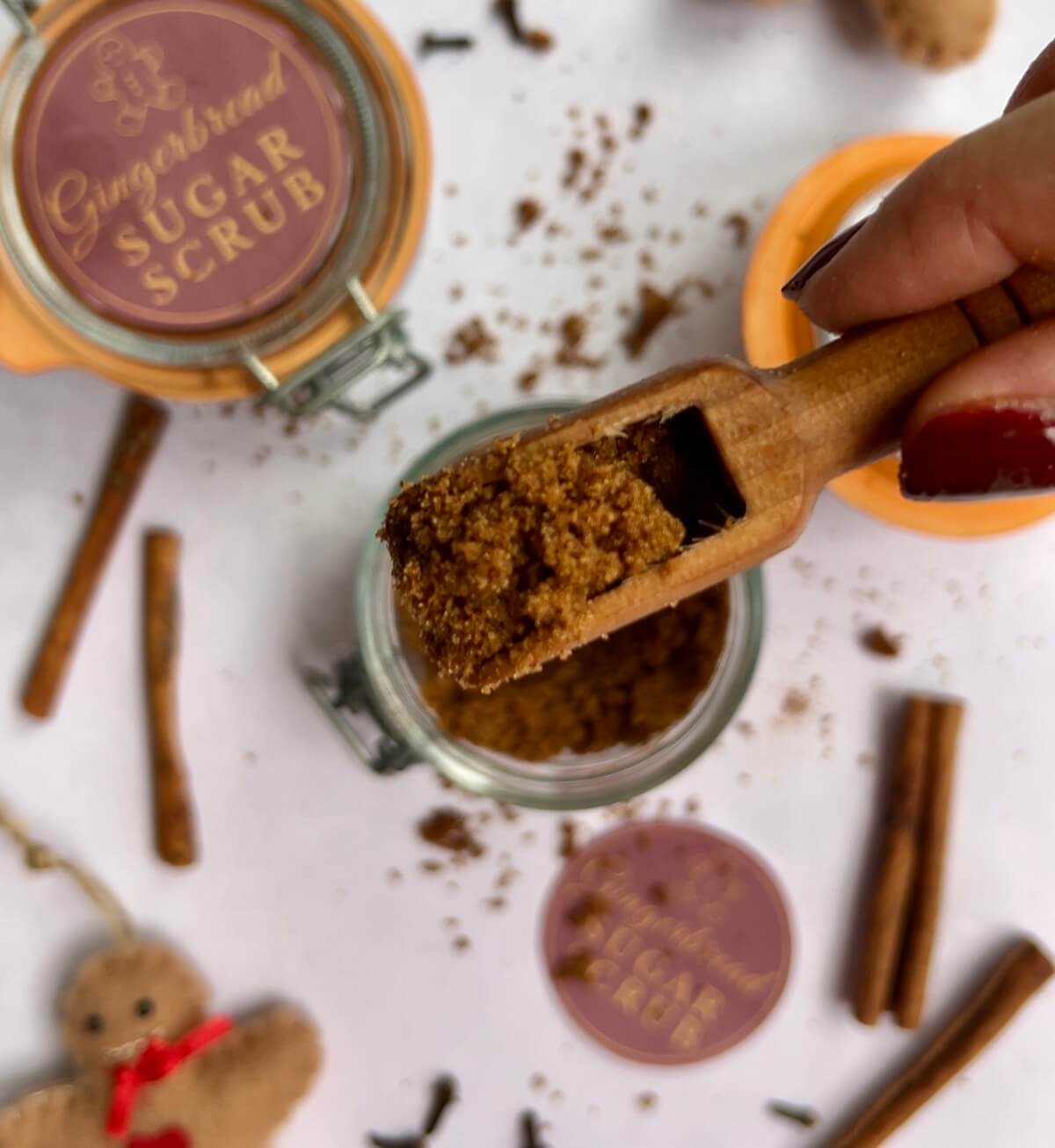 Hand holding a scoop of homemade gingerbread sugar scrub over a glass jar, with festive gingerbread men decorations and cinnamon sticks scattered around.